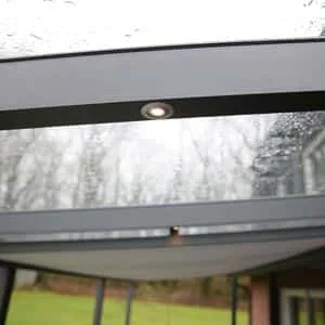 Moderne Überdachung aus Glas mit integriertem LED-Licht, fotografiert an einem regnerischen Tag, mit Blick auf einen grünen Garten im Hintergrund.