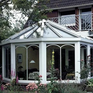 Klassischer Wintergarten in Polygonform mit weißen Rahmen und großen Glasflächen, umgeben von einem blühenden Garten.