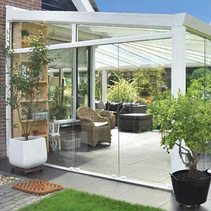 Moderner Wintergarten mit Glaswänden und weißem Rahmen, eingerichtet mit Rattanmöbeln und umgeben von grünen Pflanzen.